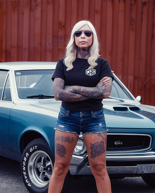 women's black factory sealed graphic tee with GHS in a shield on the left chest - woman with tattoos standing in front of a classic car with visible branding.