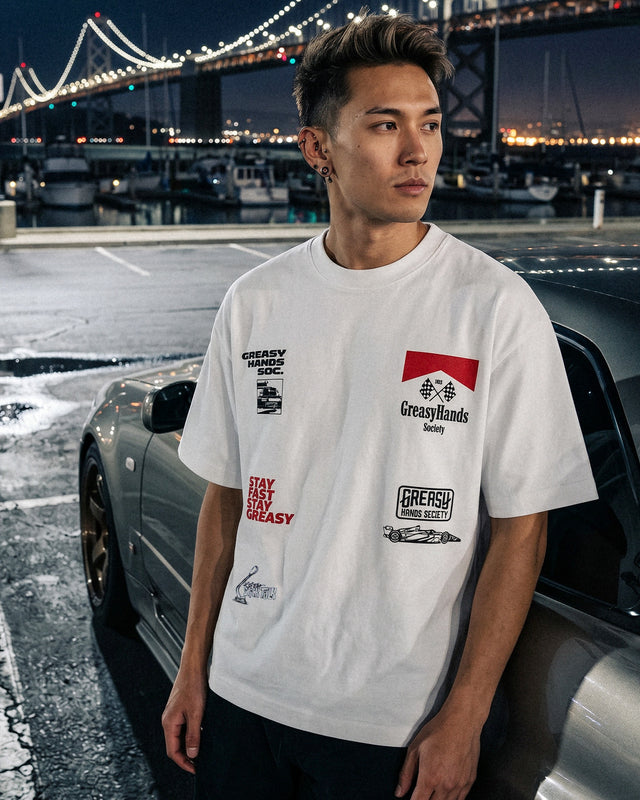Person wearing a white boxy streetwear JDM t-shirt with logos in front of a car and bridge at night.
