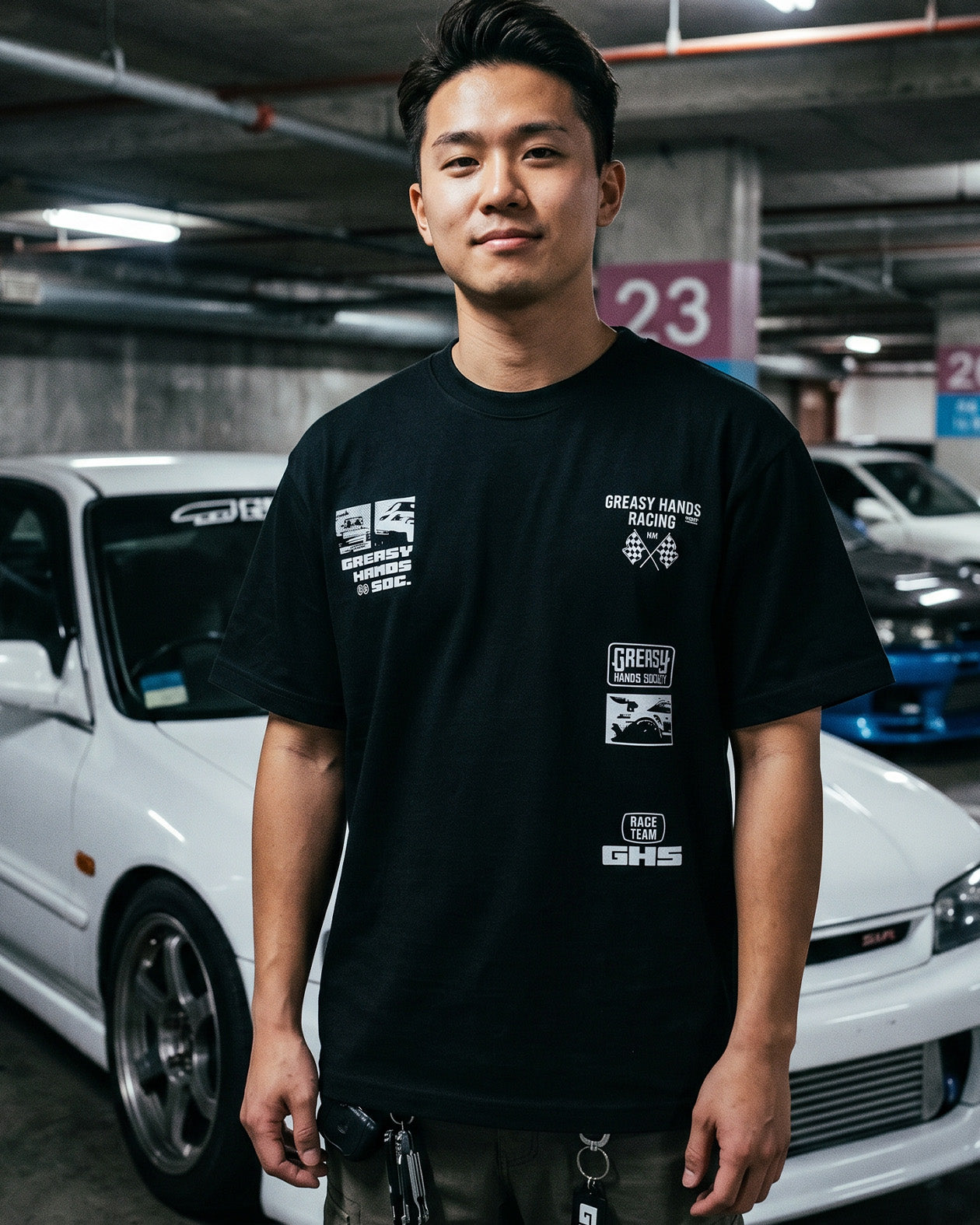 Person wearing a black t-shirt with white  logo designs  in a parking garage with cars in the background - Street Racer  graphic tee by Greasy Hands Society