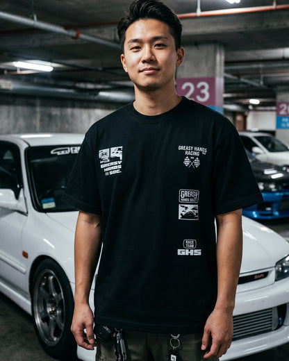 Person wearing a black t-shirt with white  logo designs  in a parking garage with cars in the background - Street Racer  graphic tee by Greasy Hands Society