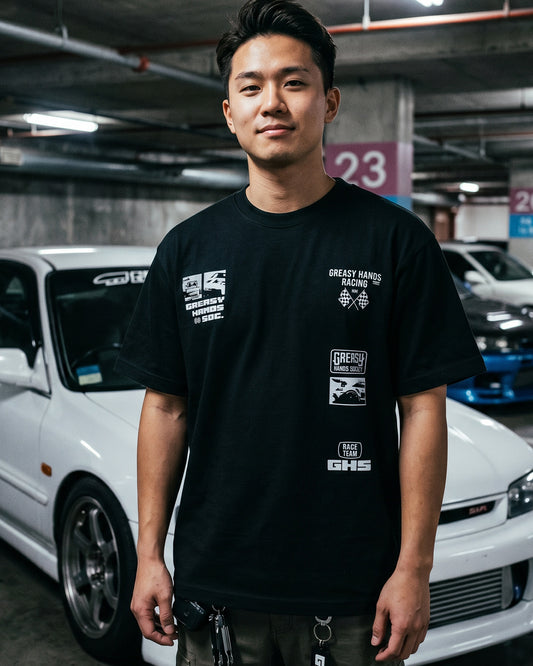 Person wearing a black t-shirt with white  logo designs  in a parking garage with cars in the background - Street Racer  graphic tee by Greasy Hands Society