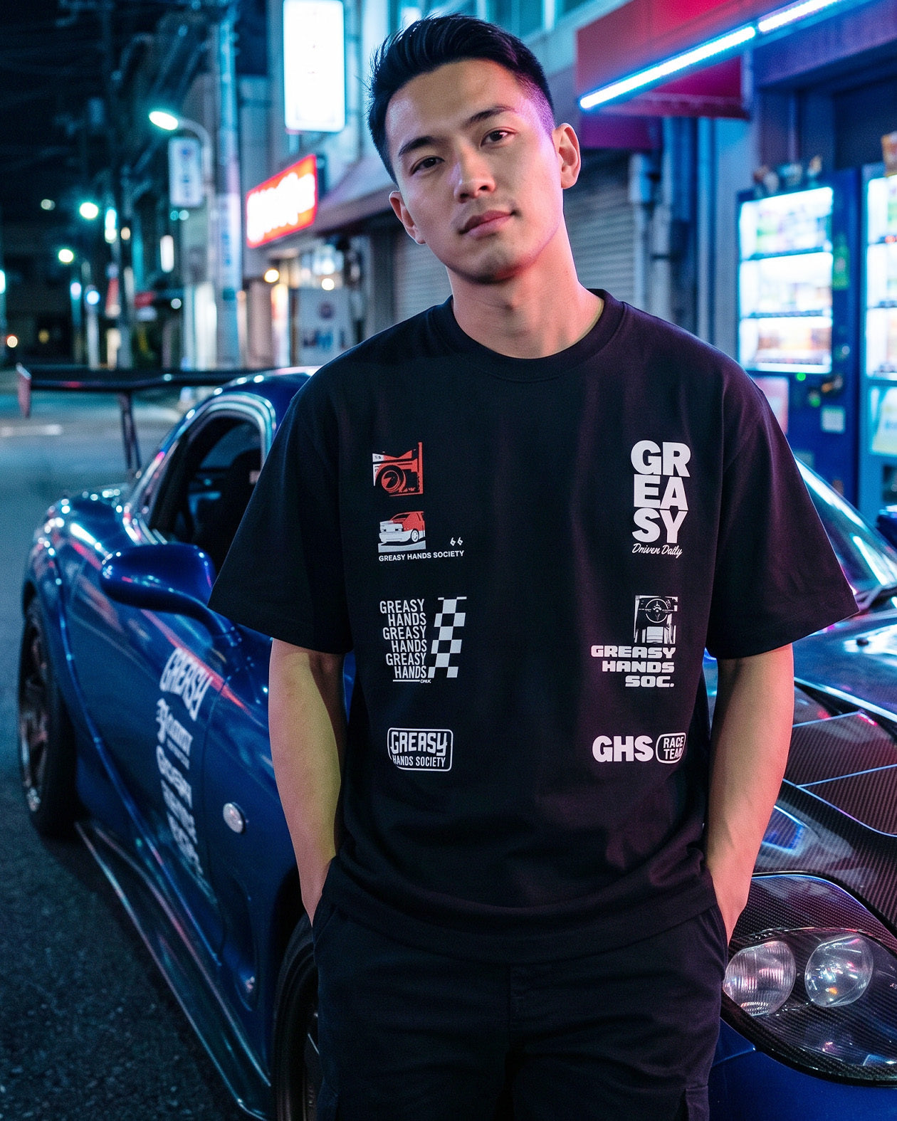 Man wearing a black t-shirt with logos standing next to a car on a street at night - JDM Super Stack Graphic Tee by Greasy Hands Society