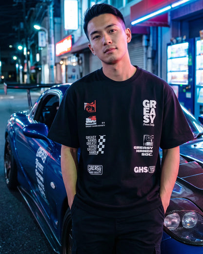 Man wearing a black t-shirt with logos standing next to a car on a street at night - JDM Super Stack Graphic Tee by Greasy Hands Society