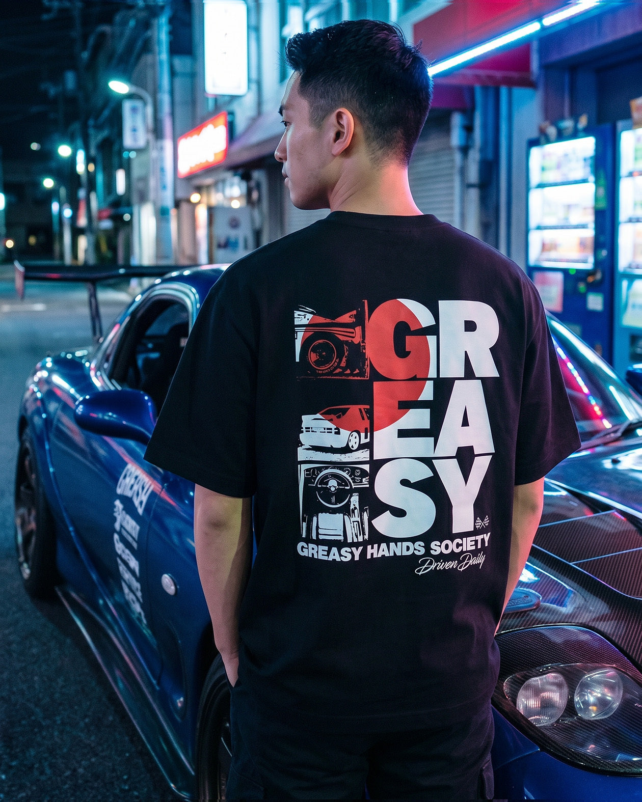 Person wearing a black t-shirt with 'Block Letters spelling "Greasy" design in white and red standing next to a car at night - The JDM Super Stack Graphic  Driven Daily Tee by Greasy Hands Society