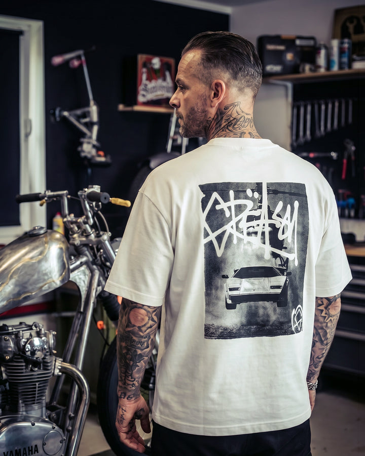 Man wearing a white t-shirt with a graphic design in a garage setting - Flying Bull White Heavyweight graphic Tee by Greasy Hands Society