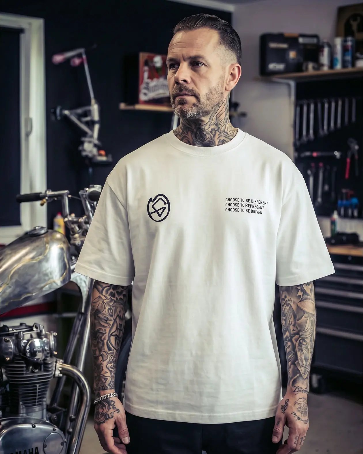 Man wearing a white t-shirt with a logo and text in a workshop setting - Flying Bull White Heavyweight graphic Tee by Greasy Hands Society