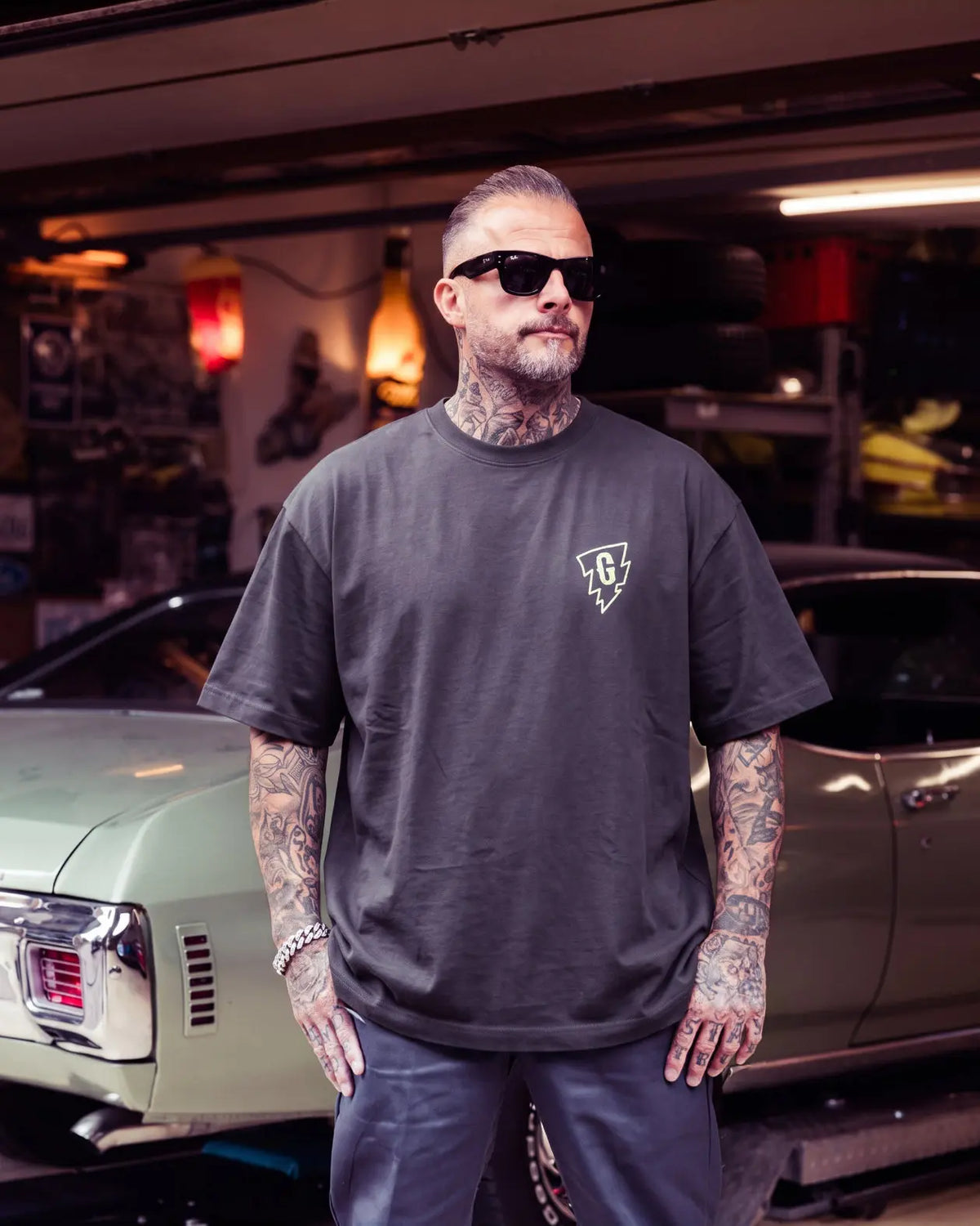 A tattooed man wearing a dark charcoal gray t-shirt with a small white chest graphic, standing in a garage with a classic car behind him - Bolt Boxy Tee by Greasy Hands Society