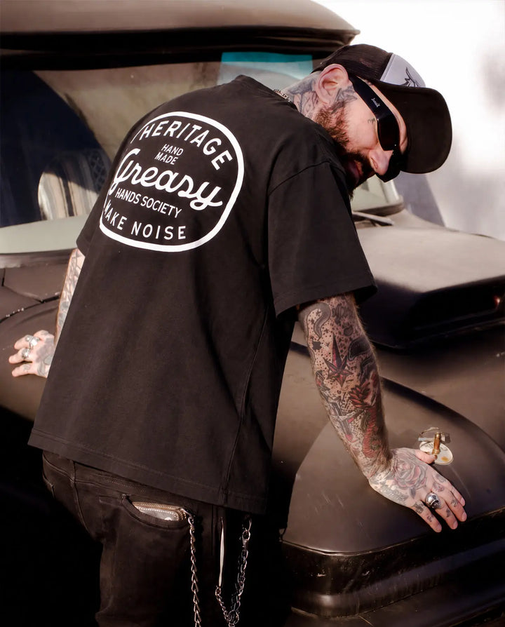 A tattooed man wearing a black t-shirt with a large white circular graphic on the back, leaning on the hood of a black car - The Heritage Boxy Graphic Tee by Greasy Hands Society
