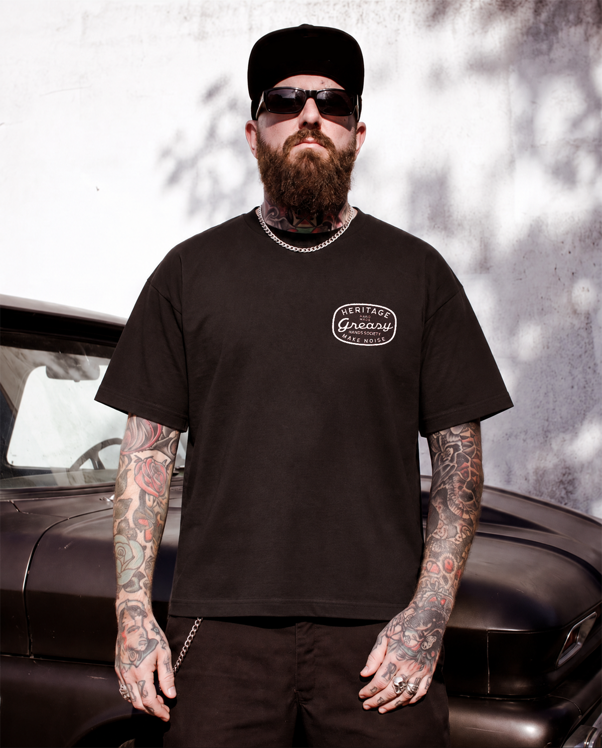 A bearded man with tattoos wearing a black t-shirt with a white 'Heritage - Greasy Hands Society - Make Noise' logo on the chest, standing in front of a vintage car - The Heritage Graphic Tee by Greasy Hands Society