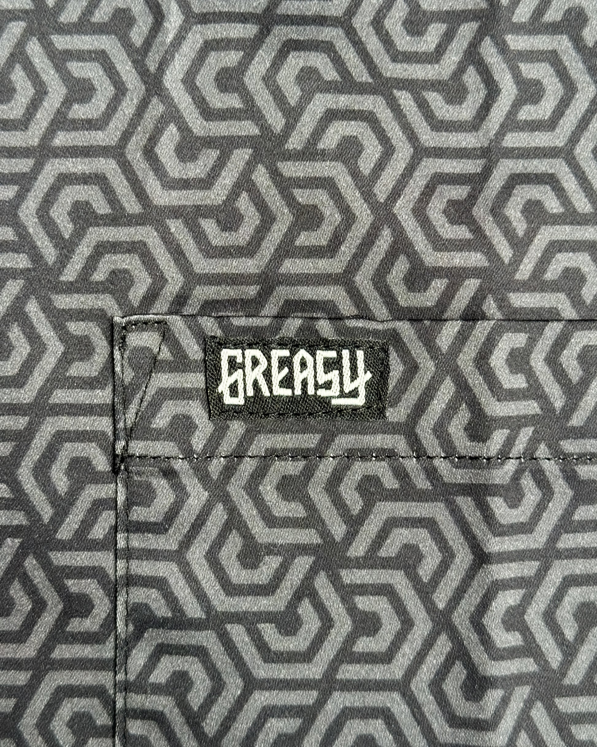 Close-up of a gray and black geometric patterned fabric with a black patch featuring the white 'GREASY' brand logo on the pocket - The Burnout Button-Up Shirt by Greasy Hands Society