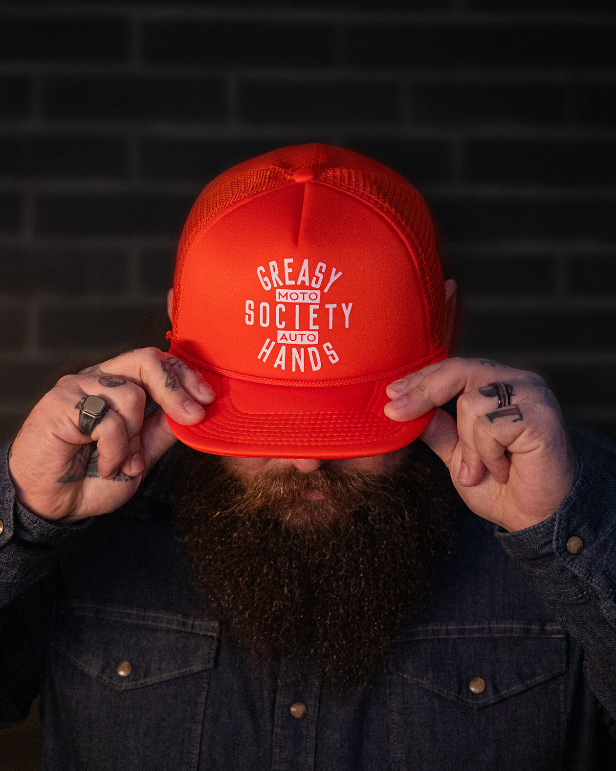 Person holding a orange cap with 'Greasy Hands Society - Moto - Auto' text against a dark brick wall.