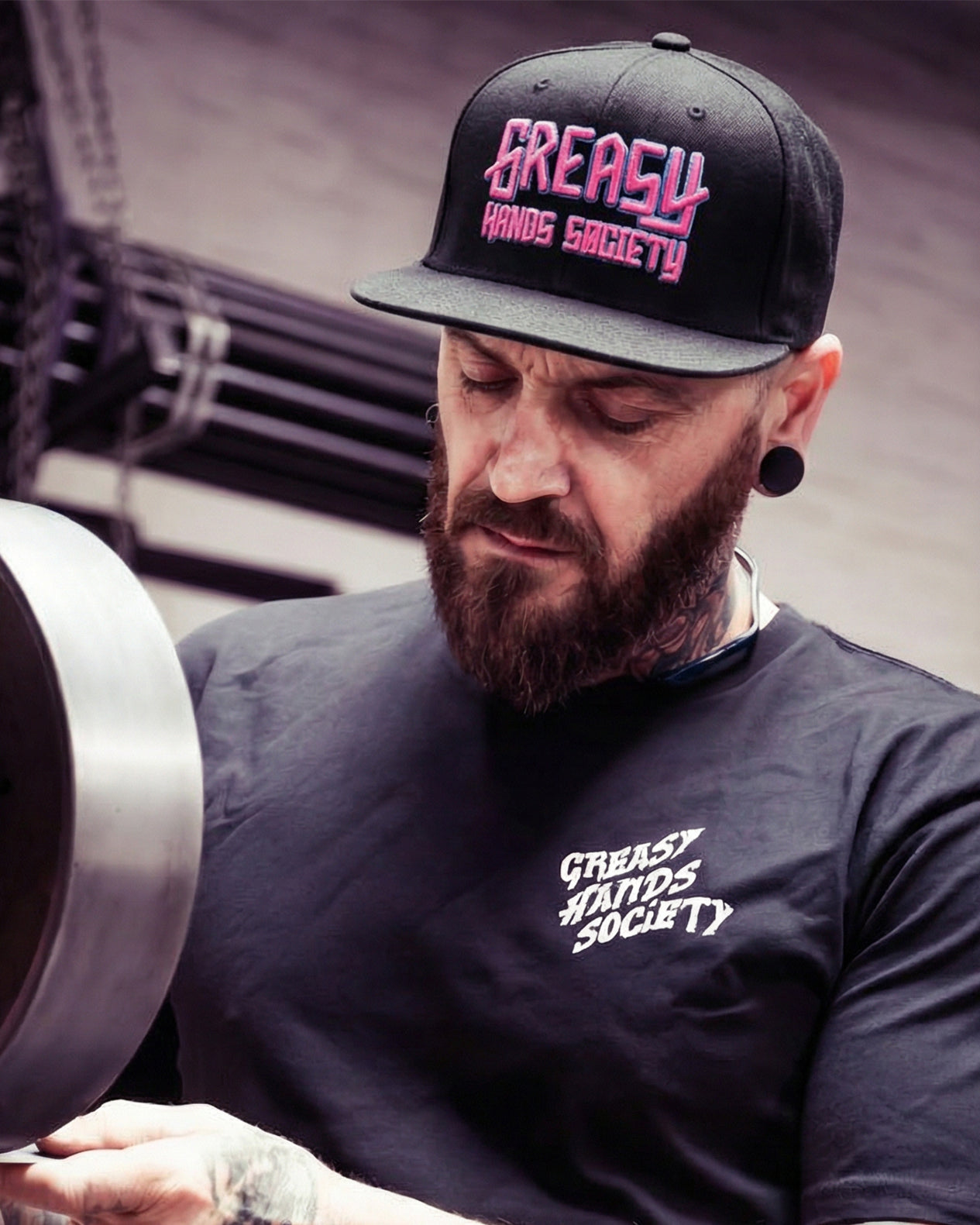 Man wearing a 'Greasy Hands Society' cap and t-shirt, holding a metal object. blue and pink pro-series black hat