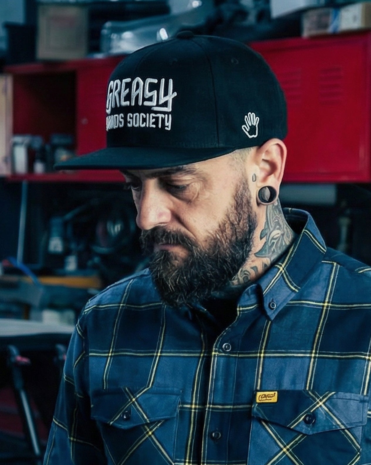 Man wearing a black cap with 'Greasy Hands Society' in white text and plaid flannel shirt in a workshop setting