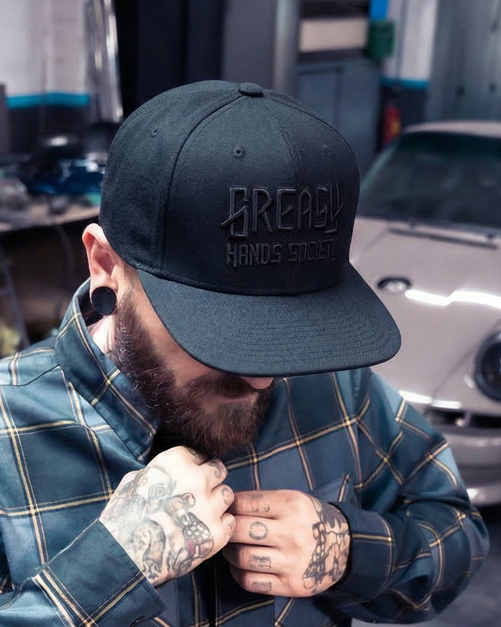 Person wearing a black cap with 'Greasy Hands Sociiety' logo in a garage setting with a car in the background.