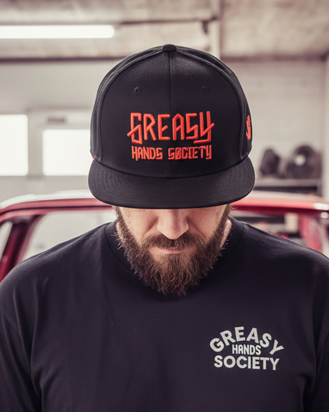 Person wearing a black cap and t-shirt with red 'Greasy Hands Society' branding in an indoor setting.