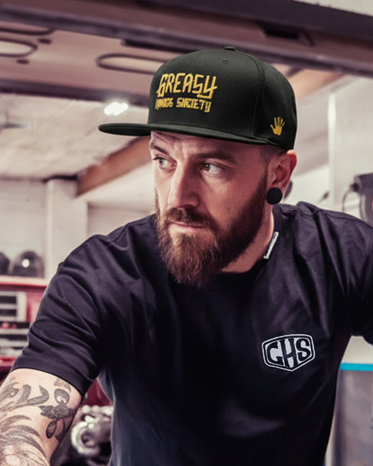 Man with tattoos wearing a black cap with gold 'greasy hands society logo' and t-shirt, working on a car in a garage.