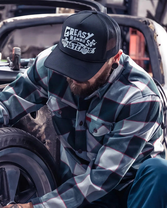 A man wearing a black trucker hat with 'Greasy Hands Society' printed in white, paired with a green and gray plaid flannel shirt, leaning over a car tire in a garage setting - Dallas Dragster Black/White Hat by Greasy Hands Society