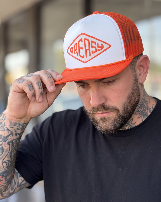 A man wearing a white and orange trucker hat with the brand name 'GREASY' printed on the front, holding the brim with his hand. The hat features a white front panel, orange mesh back, and orange brim - Cross the Line Orange/White Hat by Greasy Hands Society