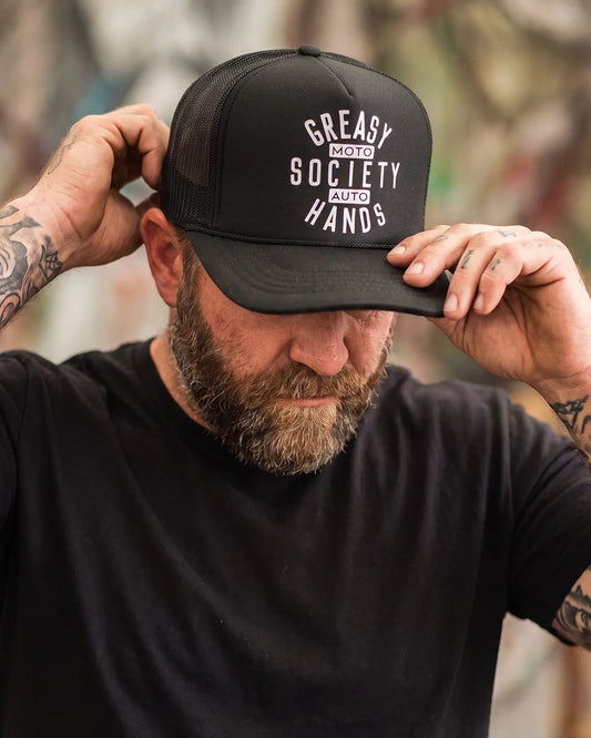 A man wearing a black trucker hat with white text reading 'Greasy Hands Society - Moto - Auto' and adjusting the brim, shown in a real-world blurred environment - Black Station Hat by Greasy Hands Society