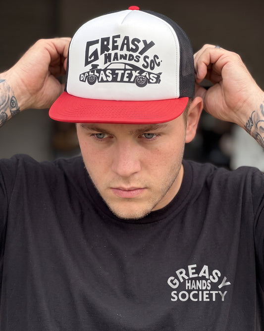 A man wearing a red and white trucker hat with 'Greasy Hands So' Las Vegas' printed on the front, adjusting the brim with both hands. He is wearing a black t-shirt with a matching white graphic on the chest - Dallas Dragster Red/White/Black Hat by Greasy Hands Society