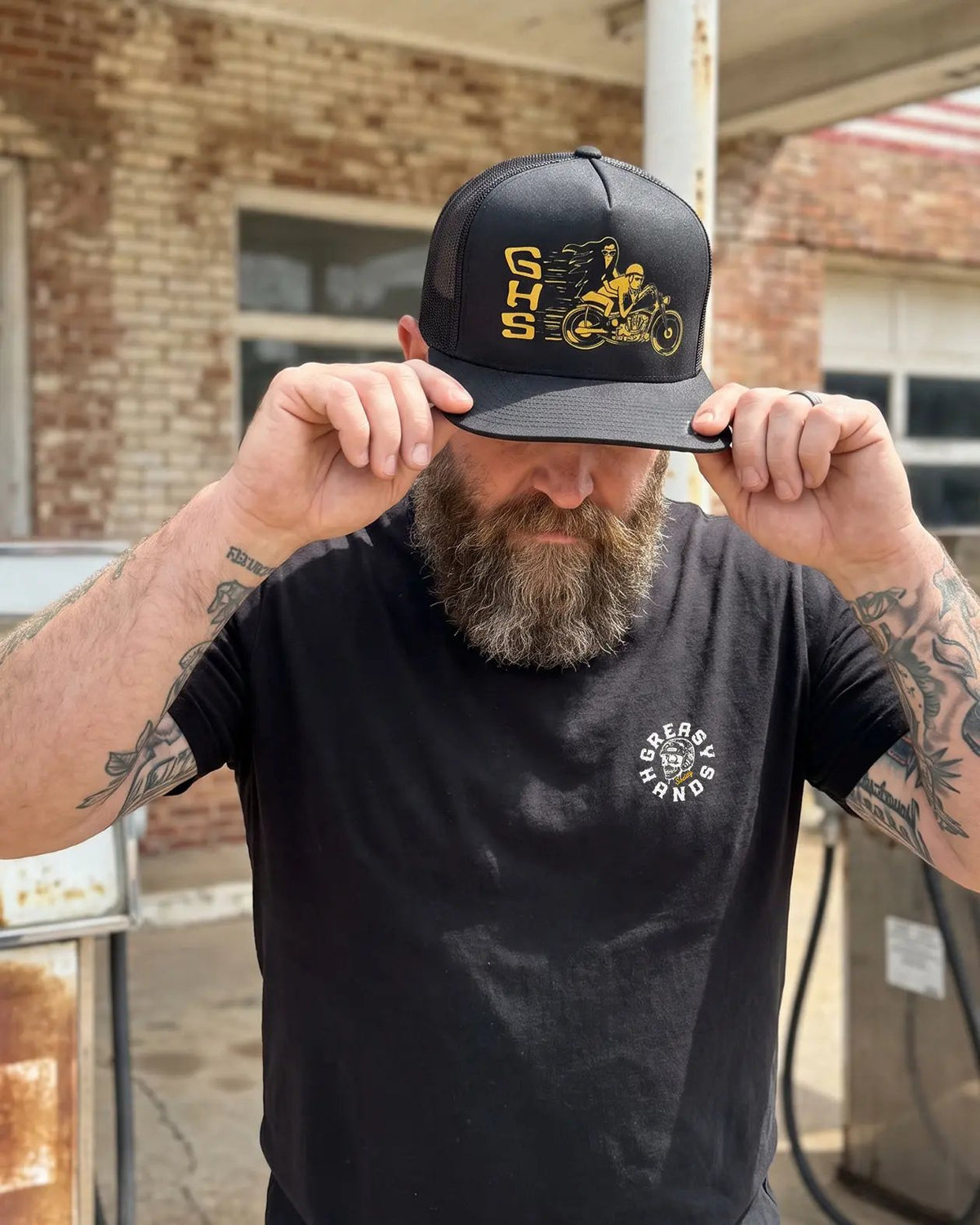 A man with a beard and tattoos wearing a black snapback cap with yellow 'CHS' lettering and a motorcycle graphic, holding the brim down over his eyes. He is wearing a black t-shirt with a white circular logo, set against a brick building background - Degenerates Hat by Greasy Hands Society