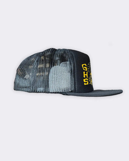 A black trucker hat with a black and gray camouflage mesh back and a black flat brim featuring yellow 'GHS' lettering on the side, set against a light gray background - Degenerates Hat by Greasy Hands Society