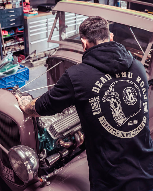 A man viewed from behind wearing a black hoodie with a large graphic on the back, leaning over the open hood of a vintage car in a garage workshop - Dead End Road Hoodie by Greasy Hands Society