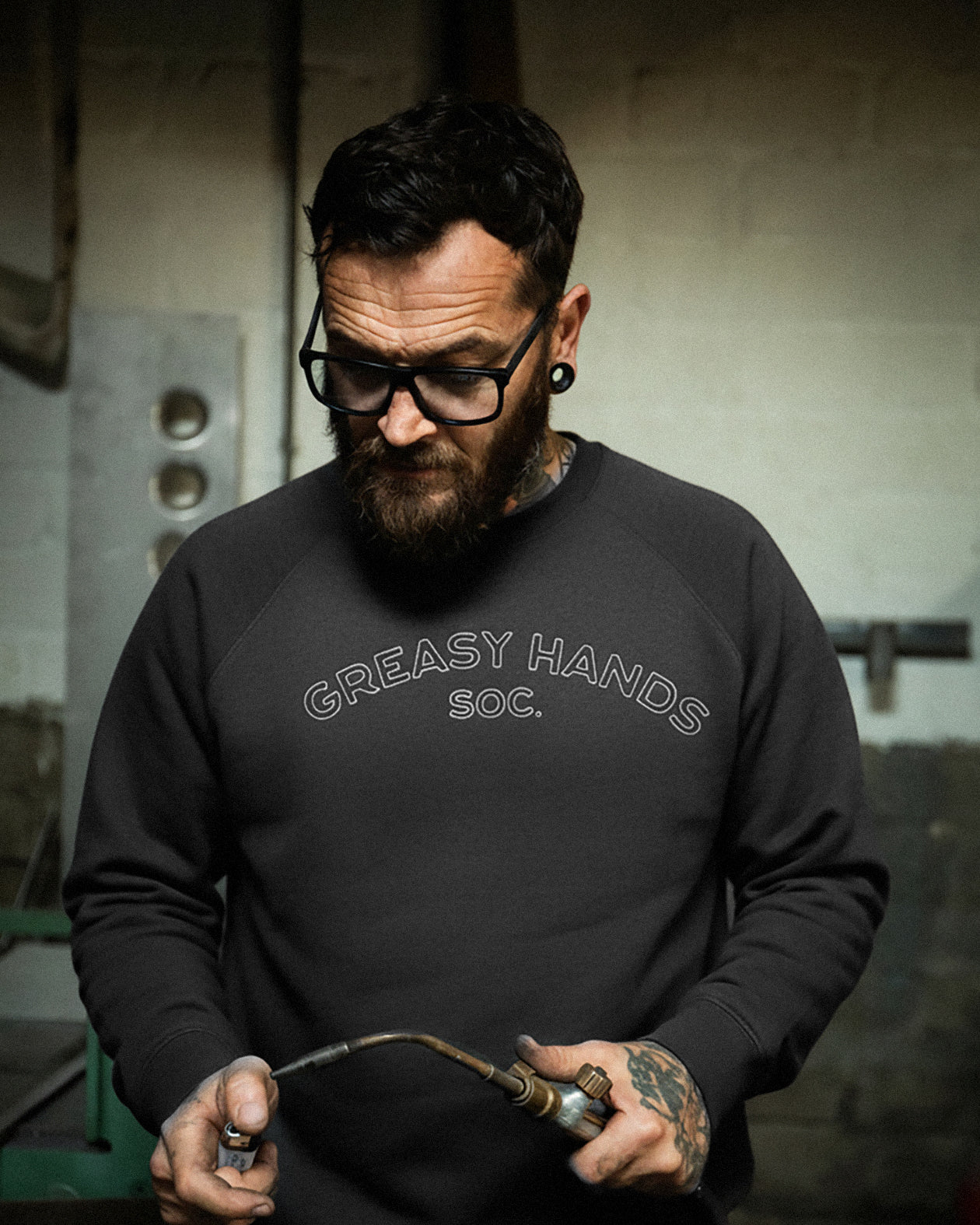 Man wearing a gray 'Greasy Hands Soc.' sweatshirt called the Joe black crew sweater working on a tool. 