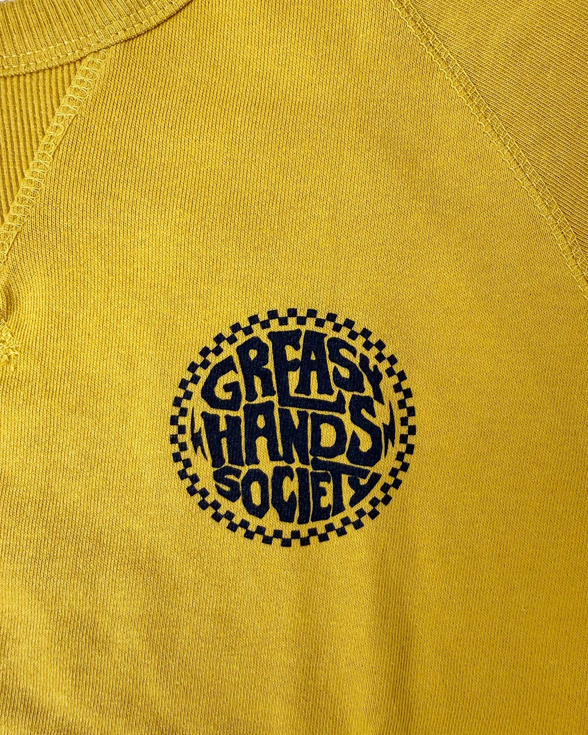 Close-up of a yellow raglan-style sweater with a circular black graphic on the chest reading 'GREASY HANDS SOCIETY' inside a checkered circle - Slicks Crew Sweatshirt by Greasy Hands Society