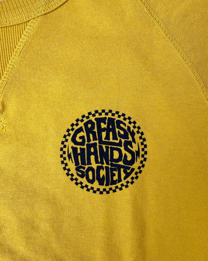Close-up of a yellow raglan-style sweater with a circular black graphic on the chest reading 'GREASY HANDS SOCIETY' inside a checkered circle - Slicks Crew Sweatshirt by Greasy Hands Society