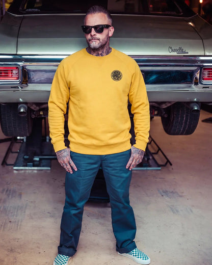 A man wearing a mustard yellow crewneck sweatshirt with a circular black graphic on the left chest, paired with dark teal pants and checkered sneakers, standing in front of a vintage car in a garage - The Slicks Crew Sweatshirt by Greasy Hands Society