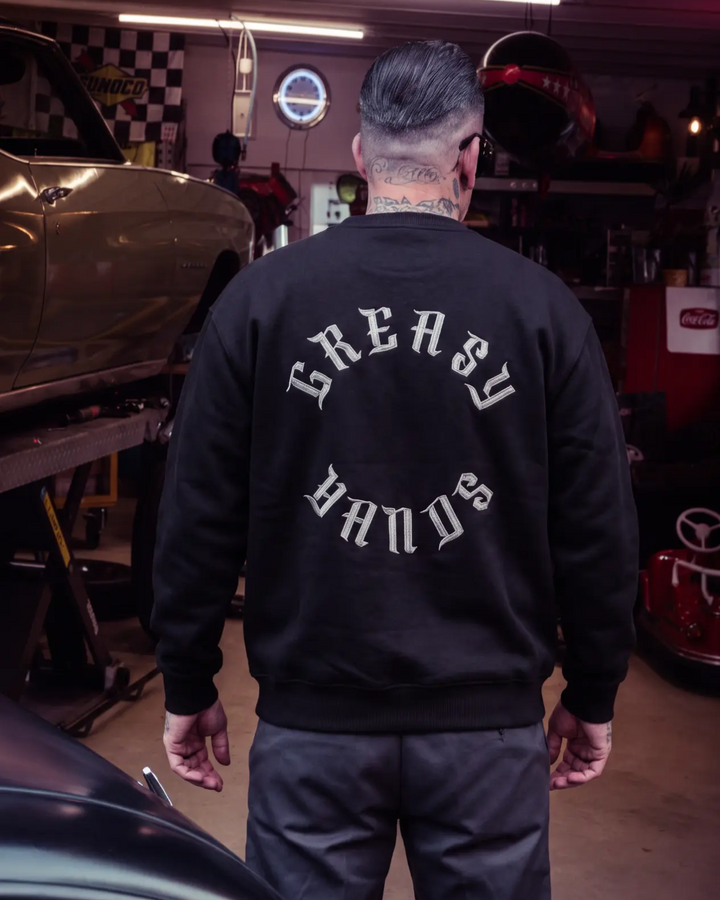 A man viewed from behind wearing a black crewneck sweatshirt with 'GREASY HANDS' printed in white gothic letters on the back, standing in a garage setting - Roller Crew Sweatshirt by Greasy Hands Society