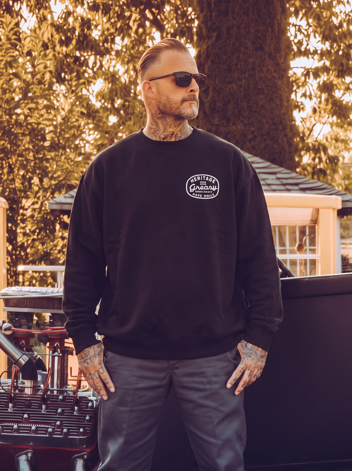 A man wearing a black crewneck sweatshirt with a white circular chest logo, dark gray pants, and white checkered sneakers, standing outdoors next to a black vehicle - The Heritage Sweatshirt by Greasy Hands Society