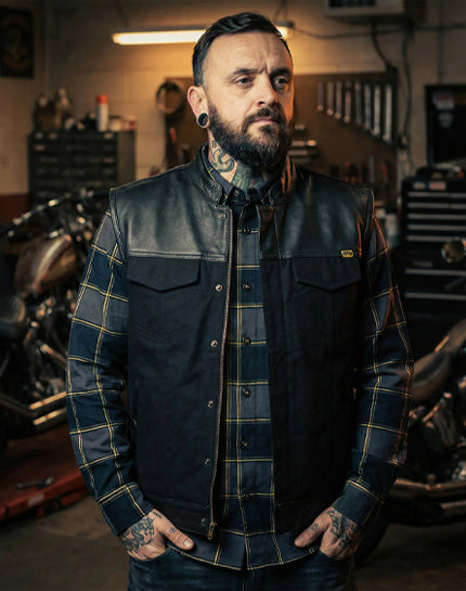 Man wearing a black leather and Canvas vest with plaid shirt underneath in a garage setting - The Jacinto Leather and Canvas Vest by Greasy Hands Society