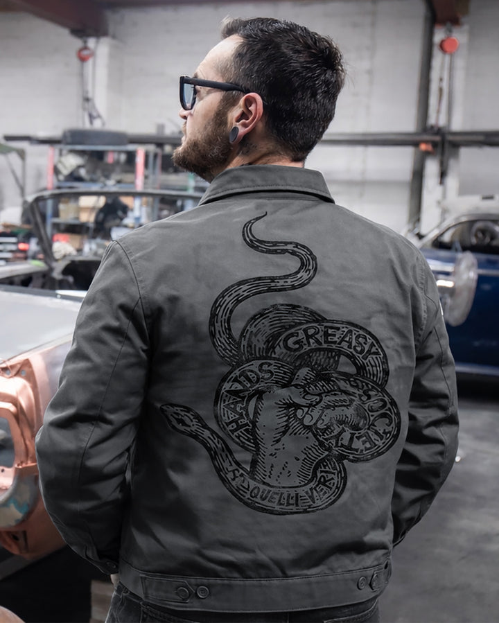 Man wearing a dark gray jacket with a snake design and text saying "Greasy Hands Society" in a garage setting 