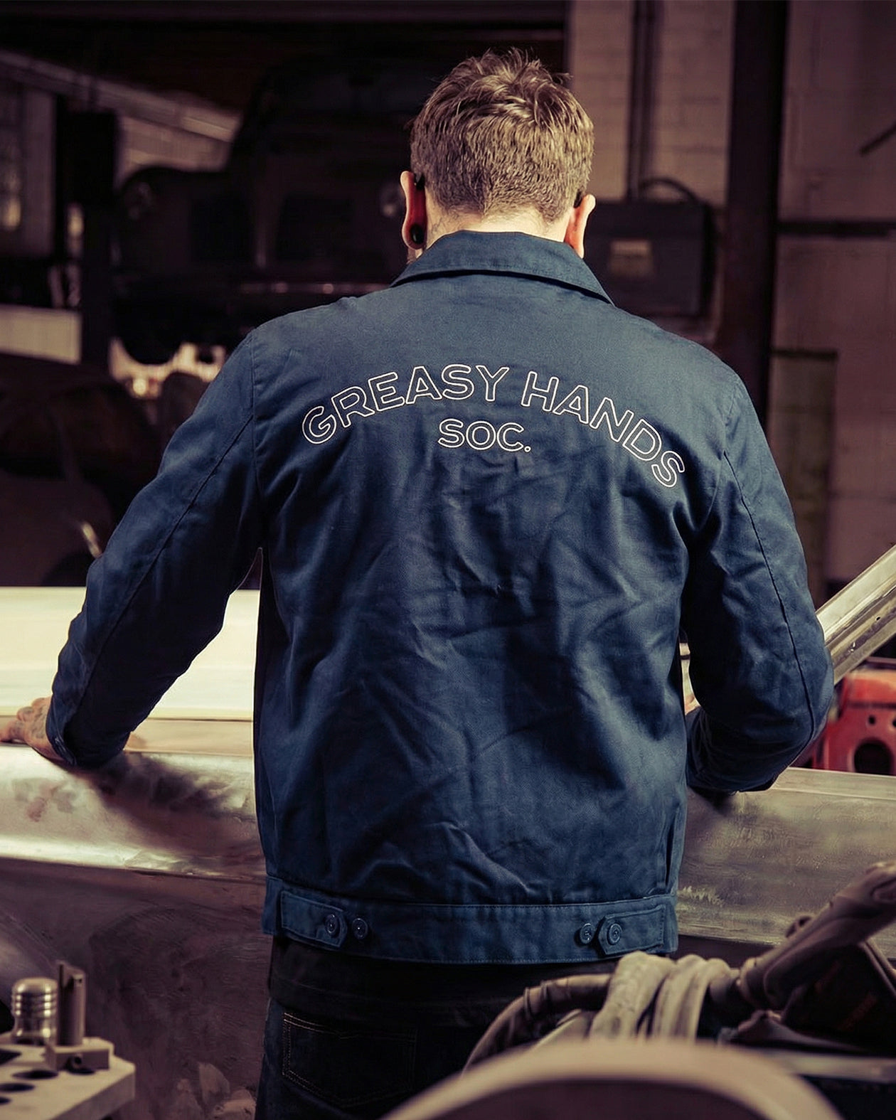 Person wearing a blue jacket with 'Greasy Hands Soc.' text in a workshop setting - Show Up Jacket by Greasy Hands Society