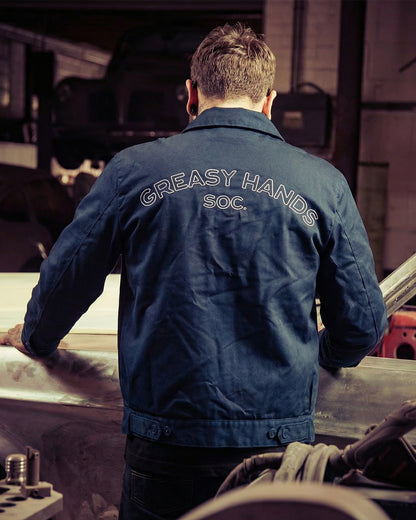 Person wearing a blue jacket with 'Greasy Hands Soc.' text in a workshop setting