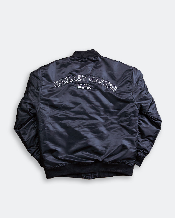 Black bomber-style jacket with 'Greasy Hands Soc' text on the back against a white background
