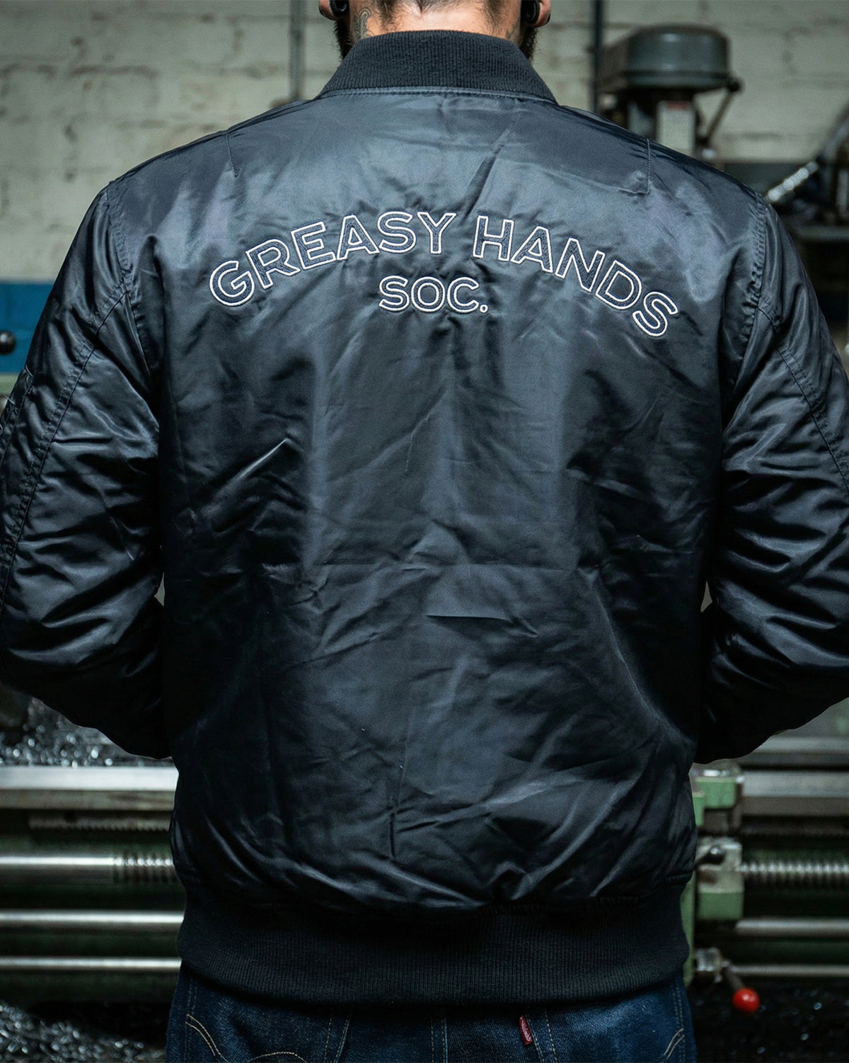 Person wearing a black flight jacket with 'Greasy Hands Soc.' text on the back, standing in an industrial setting - Men's Black Flight Jacket by Greasy Hands Society