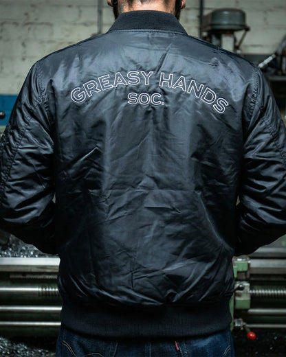 Person wearing a black flight jacket with 'Greasy Hands Soc.' text on the back, standing in an industrial setting - Men's Black Flight Jacket by Greasy Hands Society