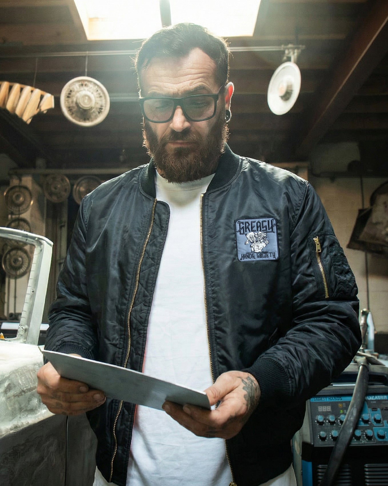 Man wearing a black flight jacket with a logo, holding a tablet in a workshop setting.