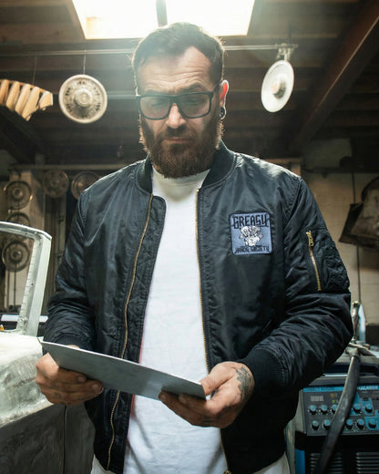 Man wearing a black flight jacket with a logo, holding a tablet in a workshop setting.