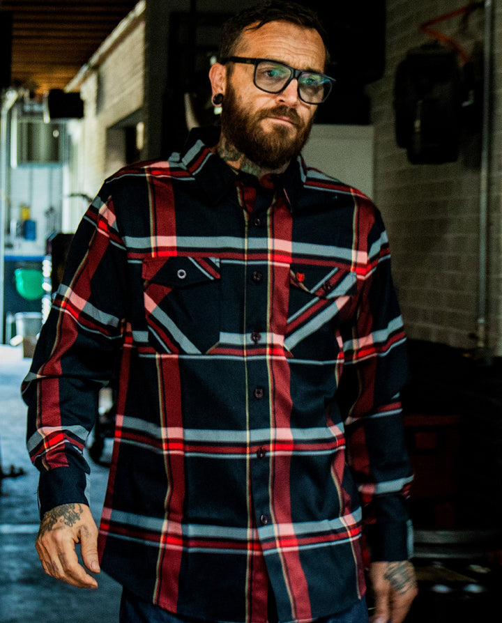 Man wearing a plaid red and black flannel shirt with tattoos on arms, standing outdoors - The Devil in the Details Flannel from Greasy Hands Society
