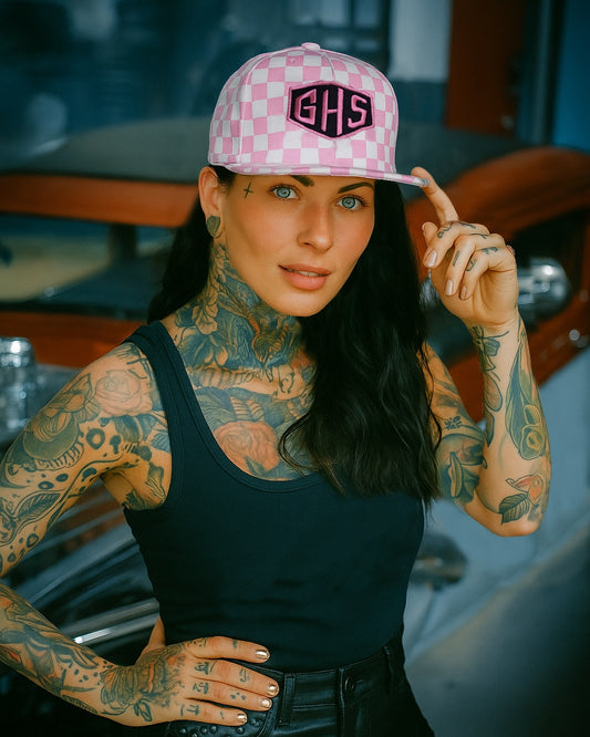 Woman wearing a pink checkerboard hat with a GHS badge, tattoos on arms and neck, black top, and leather shorts.