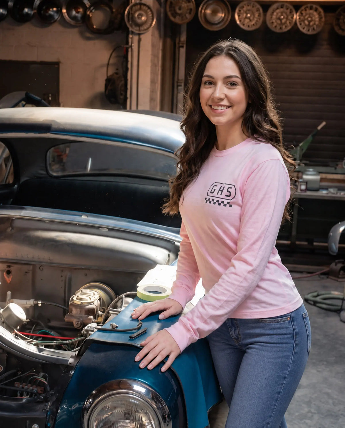a young woman standing in a workshop setting next to a vintage car that is being repaired. She is wearing the GHS engineered pink long sleeved graphic tee and jeans - GHS Engineered by Greasy Hands Society