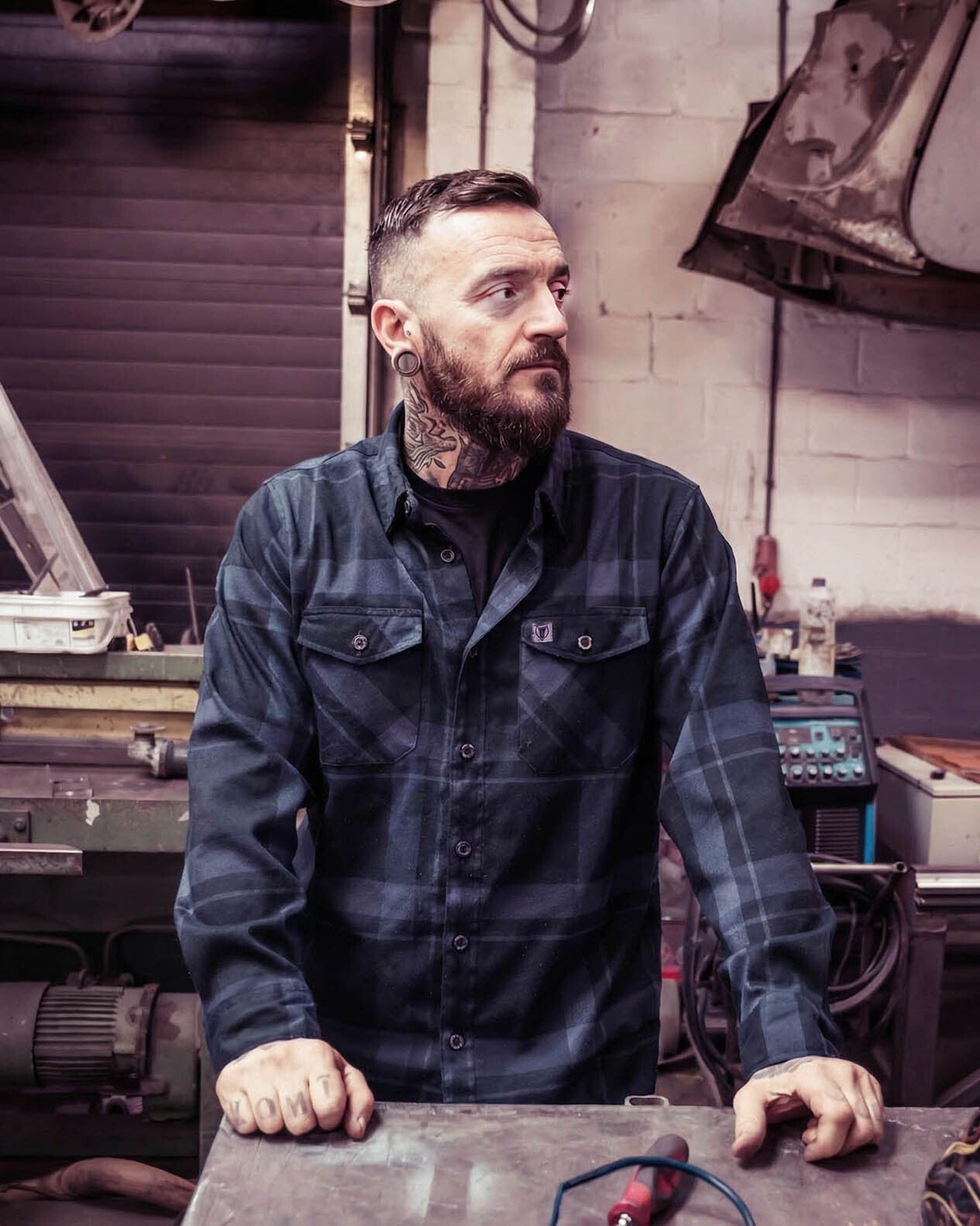 Man with tattoos and a beard wearing a black plaid shirt in a workshop setting - called the synister continental flannel