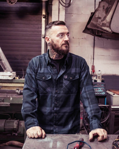 Man with tattoos and a beard wearing a black plaid shirt in a workshop setting - called the synister continental flannel