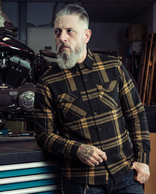Man wearing a plaid black and gold flannel shirt in a workshop setting with motorcycles - The Two-Lane Flannel from Greasy Hands Society