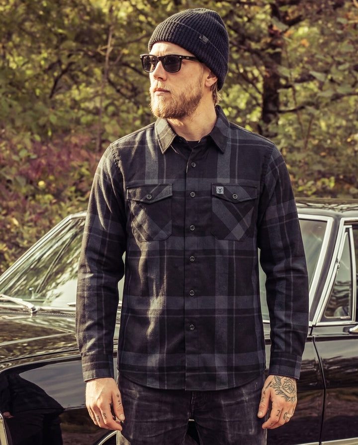 Man wearing a plaid black and gray flannel shirt and sunglasses standing next to a car with trees in the background - The Synister Flannel by Greasy Hands Society