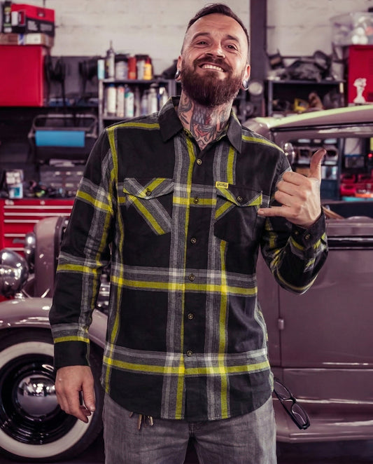 Man wearing a black and yellow plaid shirt giving a thumbs-up in a garage setting with vintage cars - The Eat Dust Flannel from Greasy Hands Society